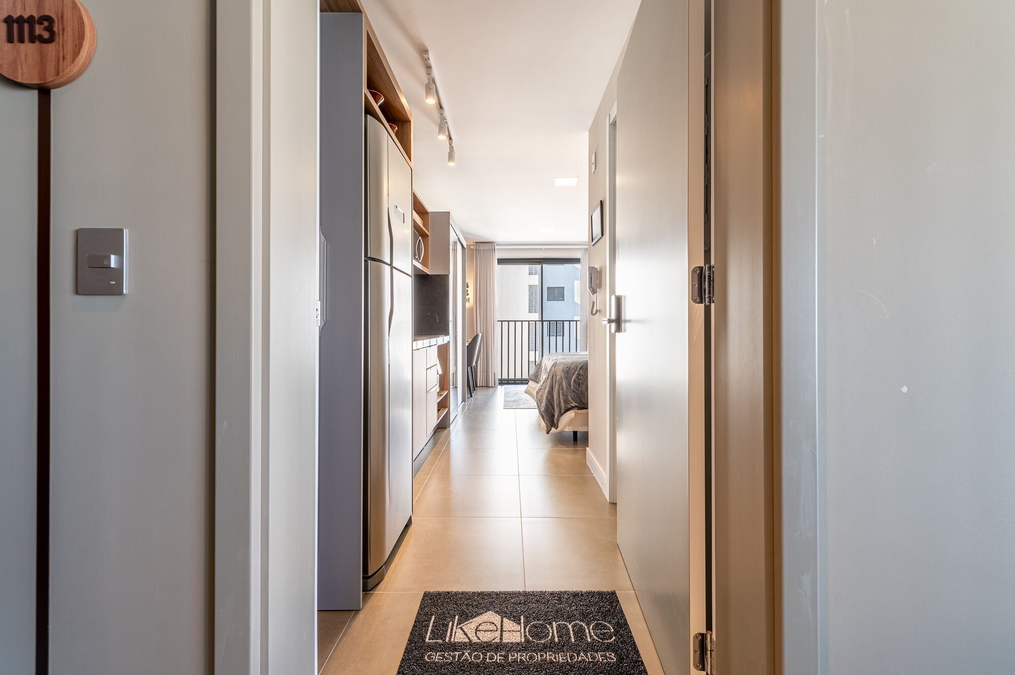 Corredor que dá acesso a um quarto com cama, escrivaninha e cadeira.