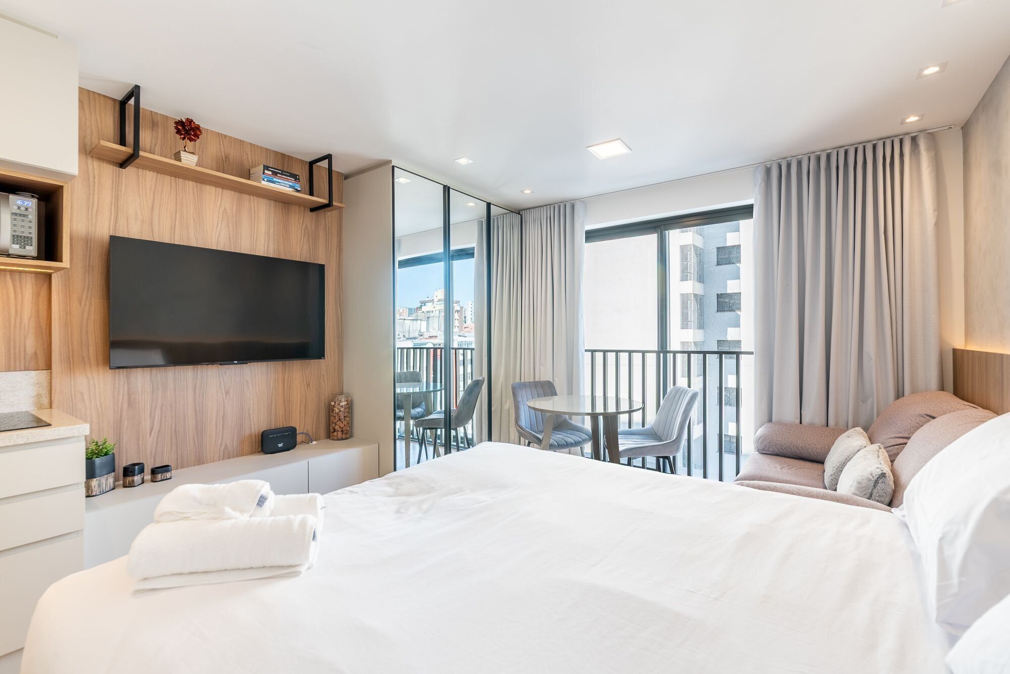 A modern hotel room with a large bed, a flat-screen TV, a balcony with a table and chairs, and a city view.