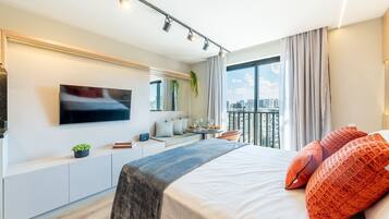 Moderni hotellihuone, jossa on sänky, televisio, oleskelualue ja parveke, jolta on näkymä kaupunkiin.