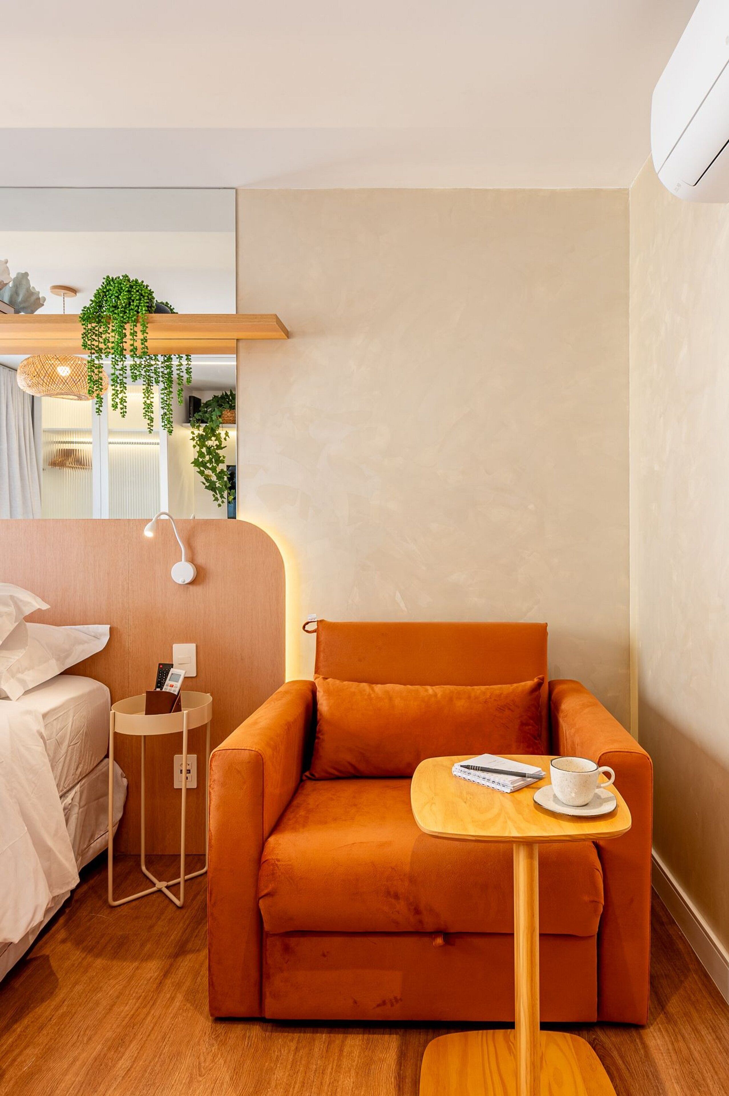 Habitación de hotel moderna con piso de madera, un sofá, una mesita con una taza y libreta, una mesita de noche con lámpara y una planta colgante.