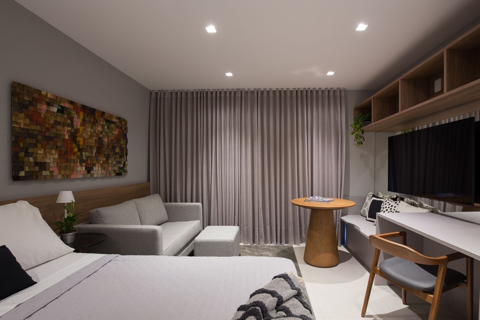 Una habitación de hotel moderna con cama, sofá, escritorio, silla, mesa redonda, televisor y cortina.