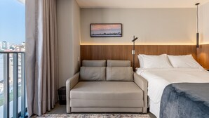 A hotel room with a bed, a sofa, a view of the city, and a cityscape painting on the wall.