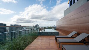 A rooftop terrace with wooden flooring, lounge chairs, and a glass railing overlooking a cityscape.