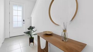 En gang med en hvid dør, et træbord med en vase og en skål, samt et stort rundt spejl på væggen.