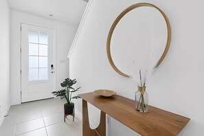 Ein Flur mit einer weißen Tür, einem Holztischchen mit einer Vase und einer Schale sowie einem großen runden Spiegel an der Wand.