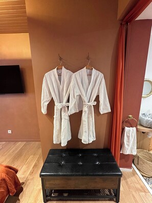 Ein Zimmer mit zwei weißen Bademänteln, die an einer Wand hängen, einer schwarzen gepolsterten Bank und einem Holzboden.