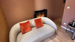 Ein Wohnzimmer mit einer Couch, zwei orangenen Kissen, einem Kamin und einem kleinen Tisch mit einem Metalltopf.