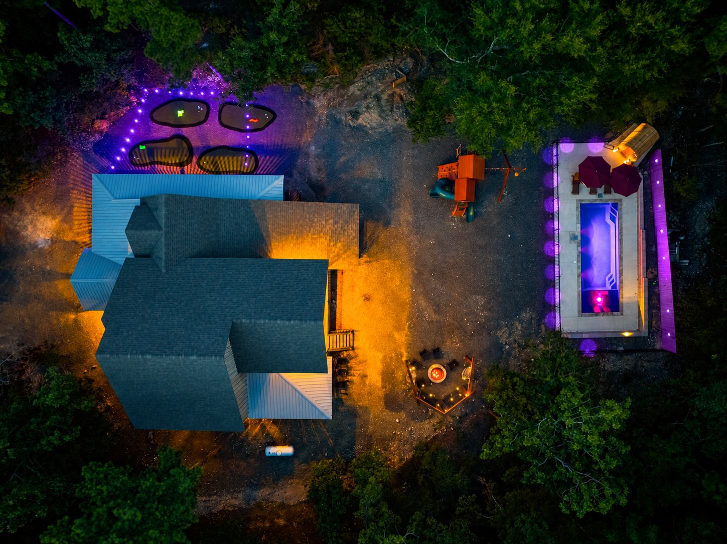 An aerial view of a property with a large building, outdoor seating area with tables and chairs, a hot tub, and a small structure with a window.
