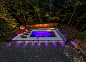 A rectangular outdoor swimming pool with a wooden deck, surrounded by a metal fence, and illuminated by purple lights.