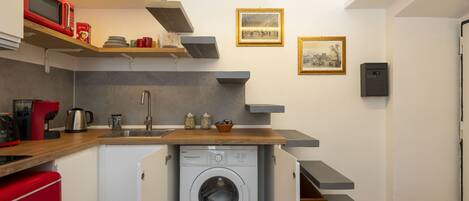 A compact kitchen with a built-in washing machine, wooden shelves, and a microwave.