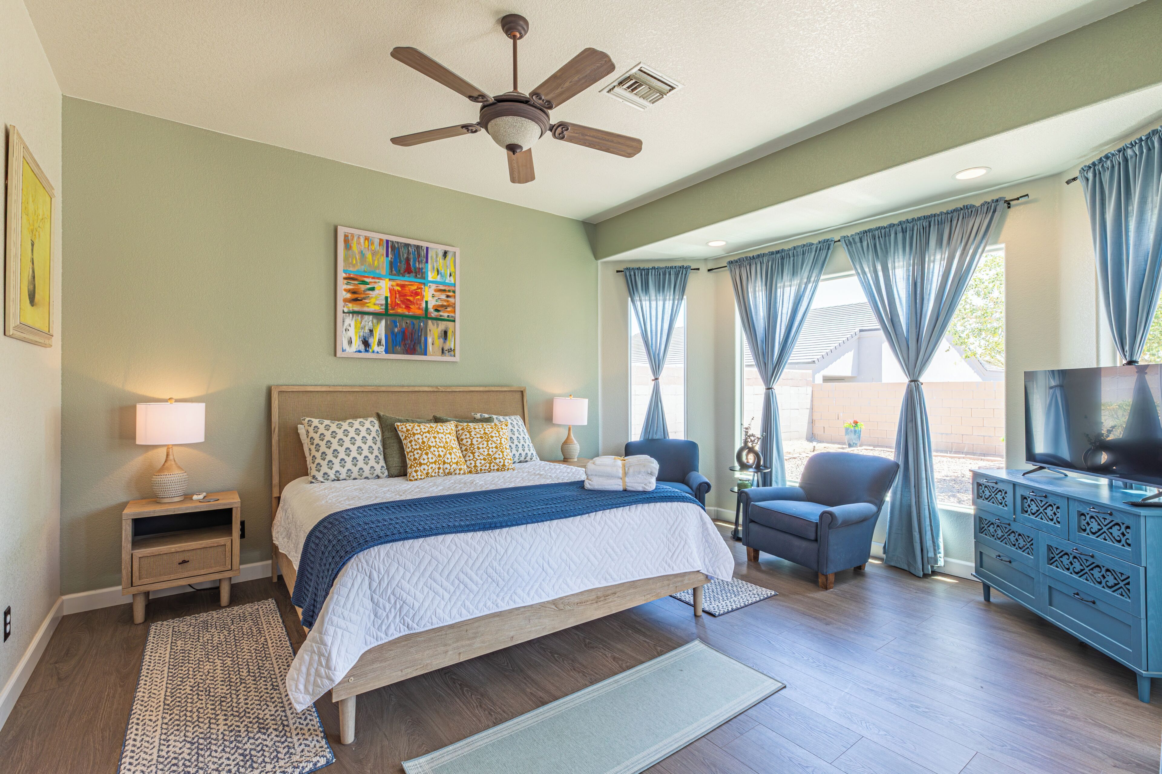 A bedroom with a bed, nightstands, a ceiling fan, a TV, a chair, a dresser, and a window with curtains.