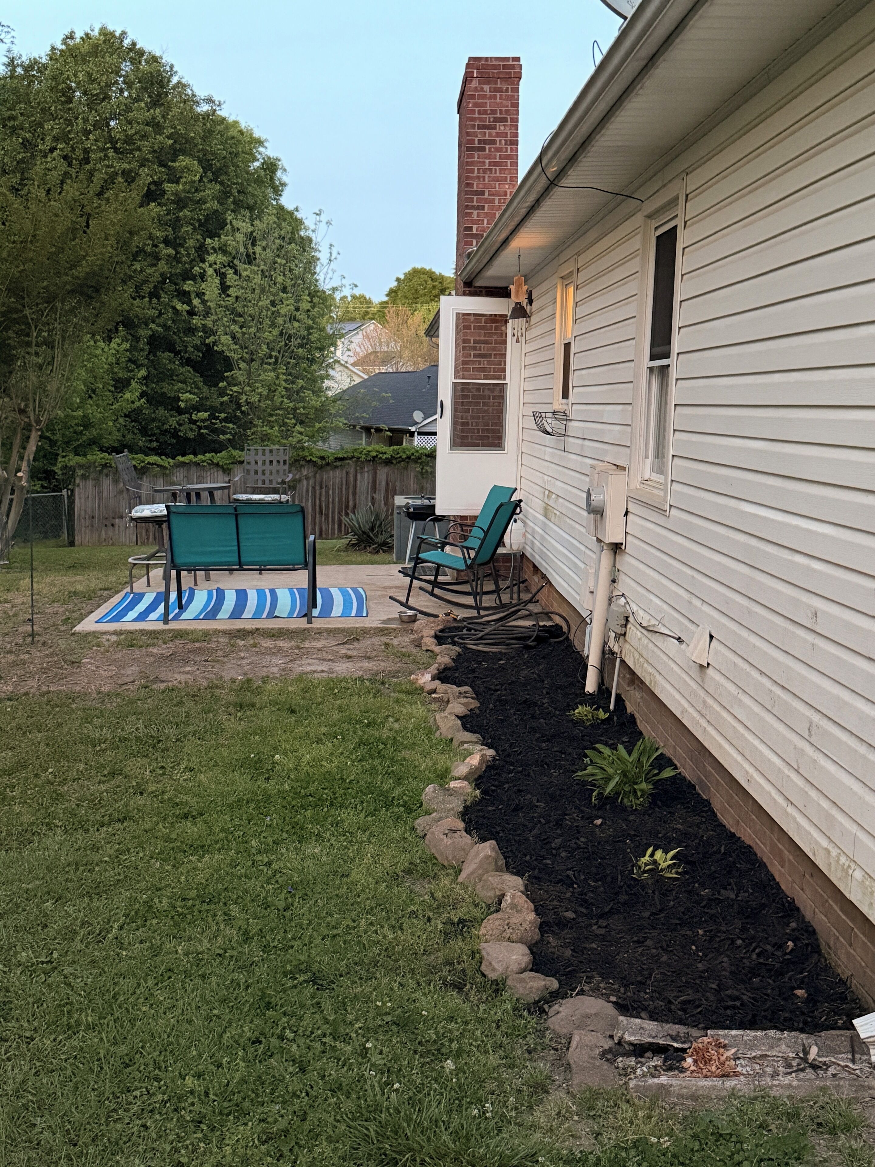 Een achtertuin met terras, tuinmeubilair, een bakstenen schoorsteen en een houten omheining.