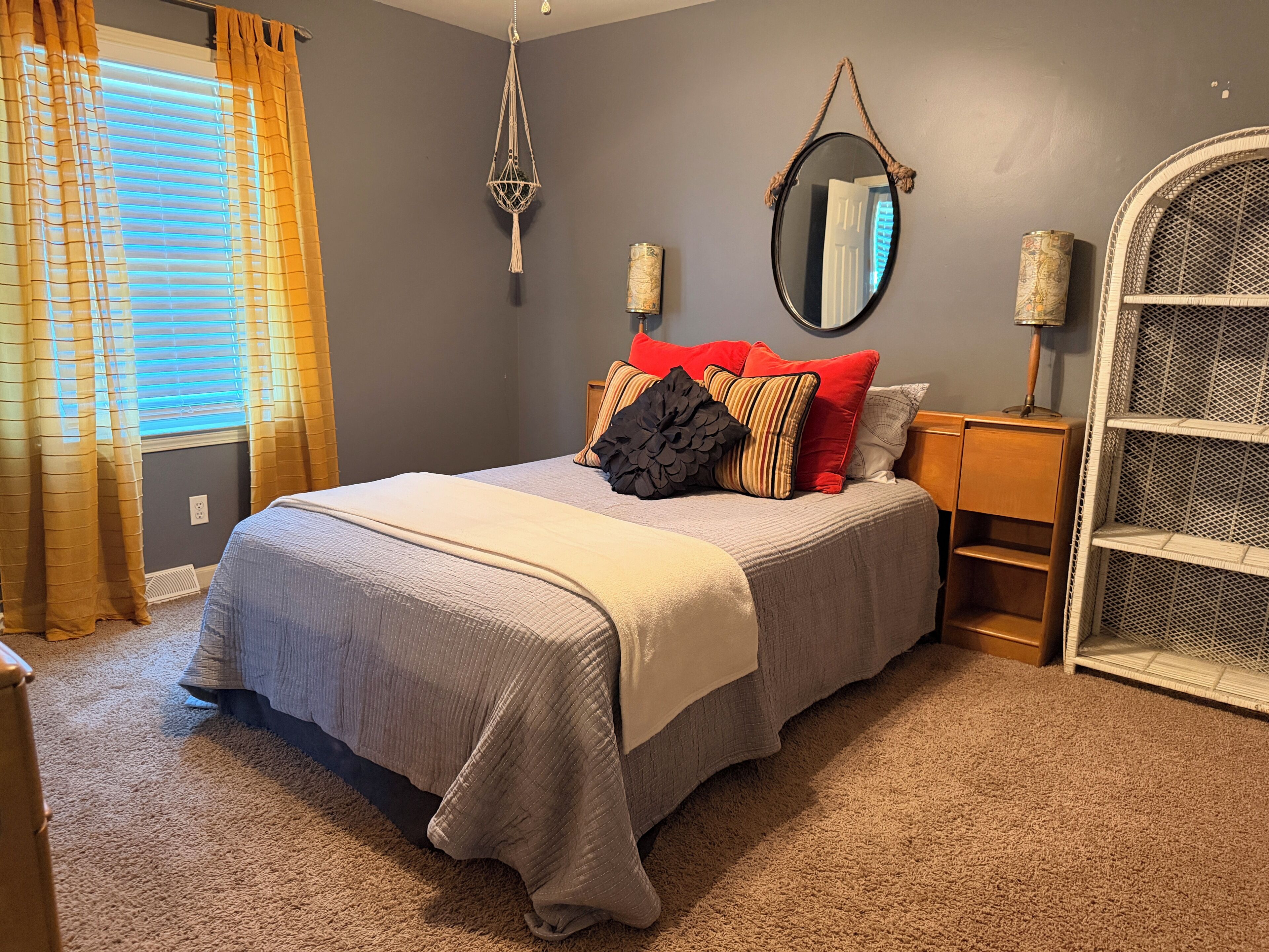 A bedroom with a bed, a nightstand, a mirror, a hanging macrame light, and a window with curtains.