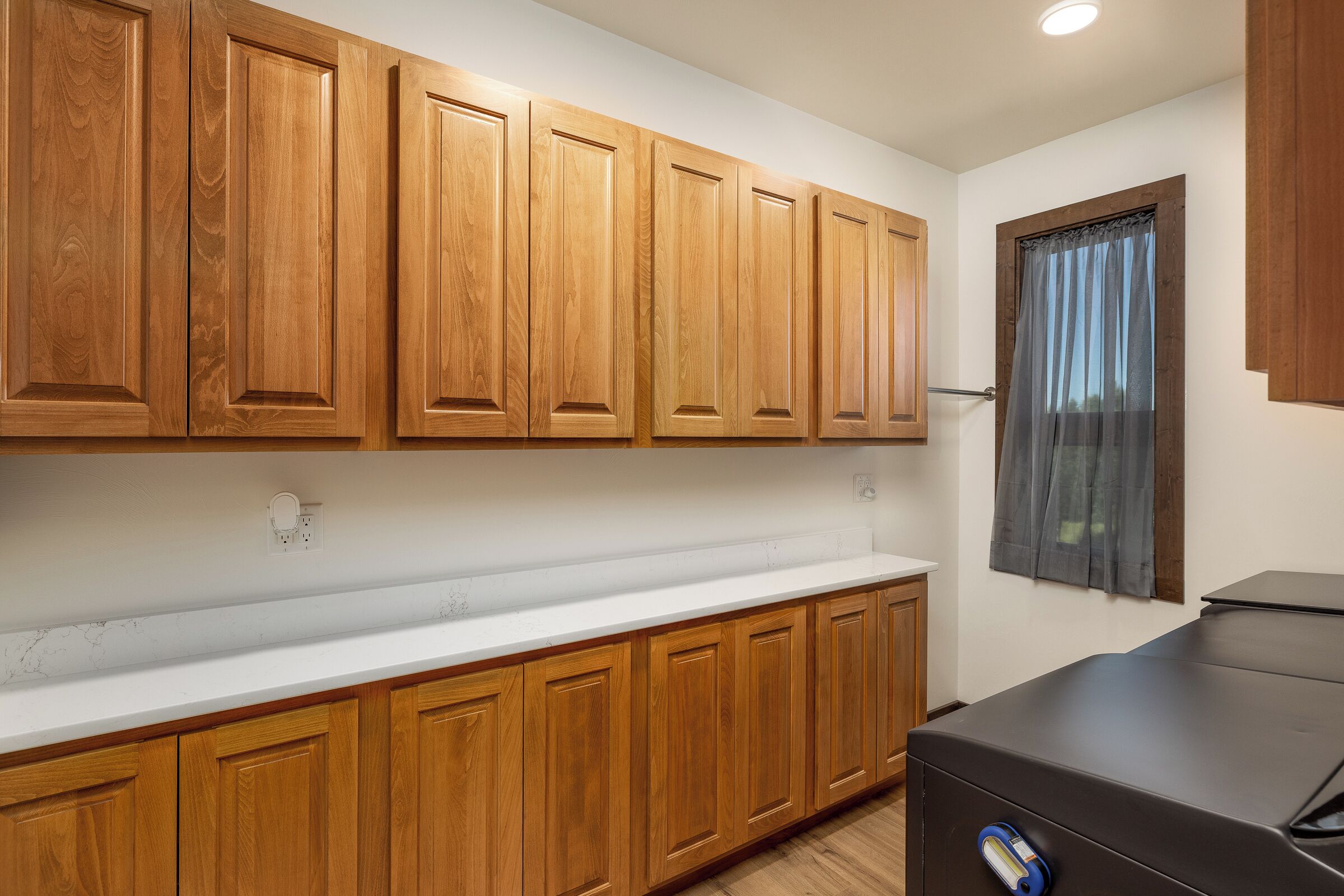 A kitchen with wooden cabinets, a white countertop, and a window with curtains.