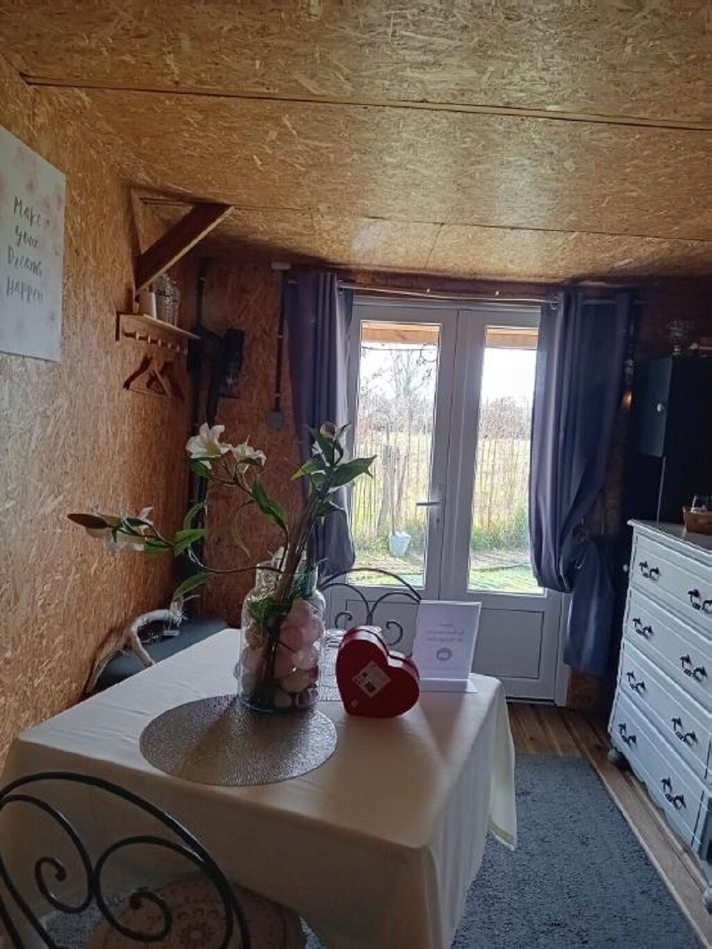Ein Zimmer mit Holzboden, einem Tisch mit weißer Tischdecke, einer Blumenvase, einem roten herzförmigen Gegenstand und einem Fenster mit Vorhängen.