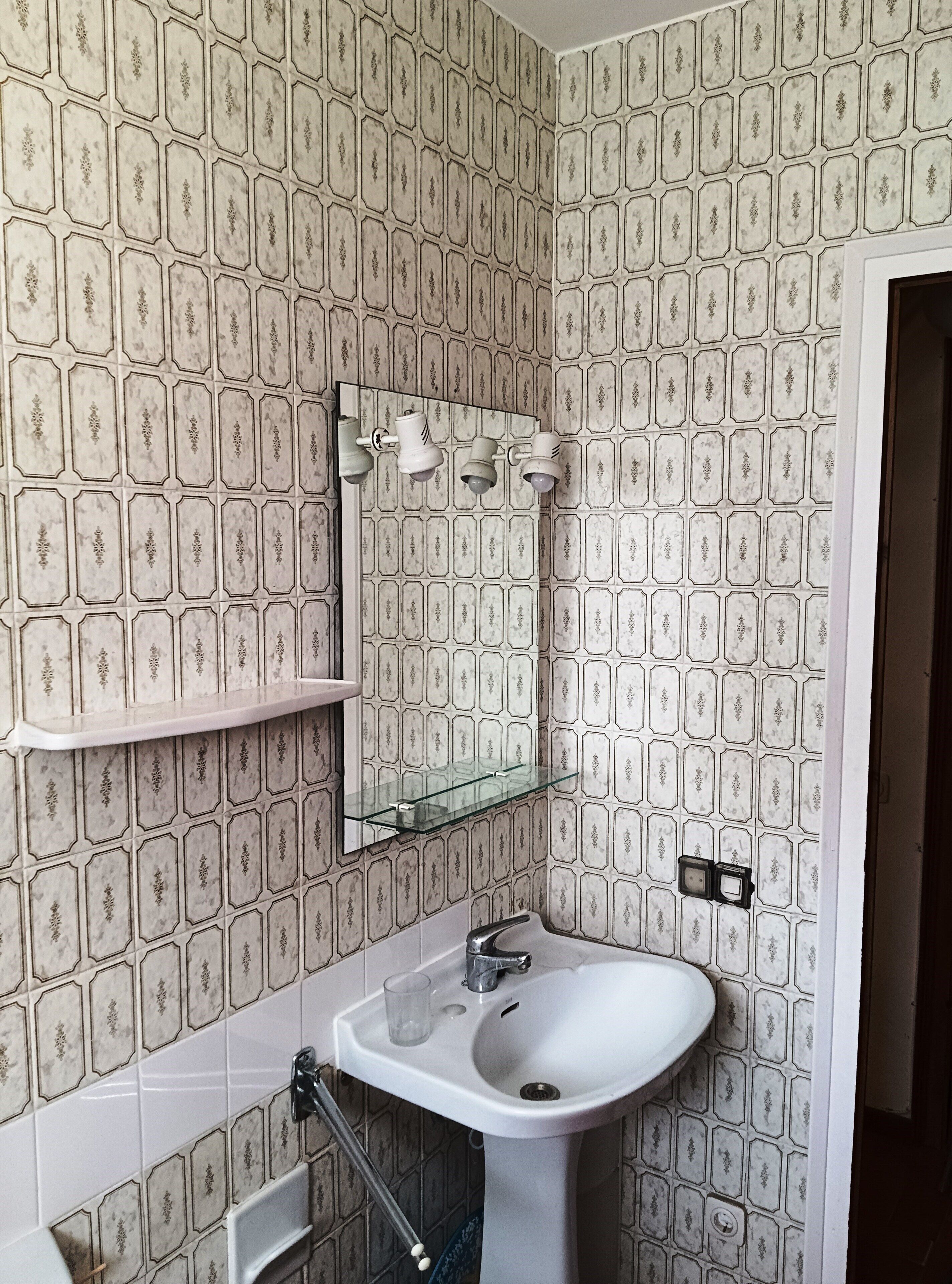 A bathroom with hexagonal patterned tiles, a white sink, a glass shelf, and a mirror with three wall-mounted lights.