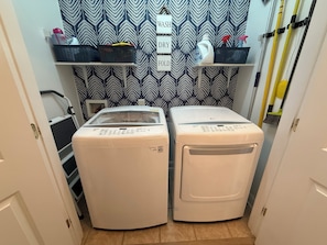 A laundry room with two front-loading washing machines, a shelf with cleaning supplies, and a patterned wallpaper. - Pink Par-Tee Villa   Luxe Beach Getaway with Golf Cart (Murrells Inlet)