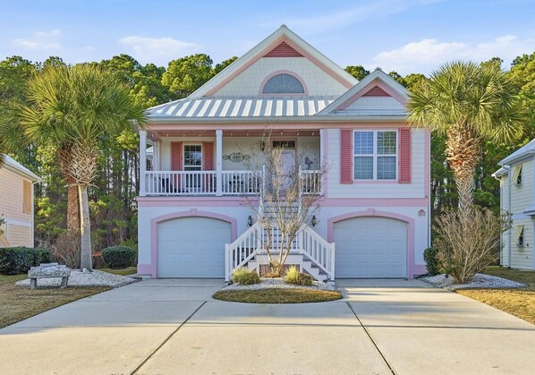 A two-story house with a front porch, a garage, and a small front yard with a palm tree. - Pink Par-Tee Villa   Luxe Beach Getaway with Golf Cart (Murrells Inlet)