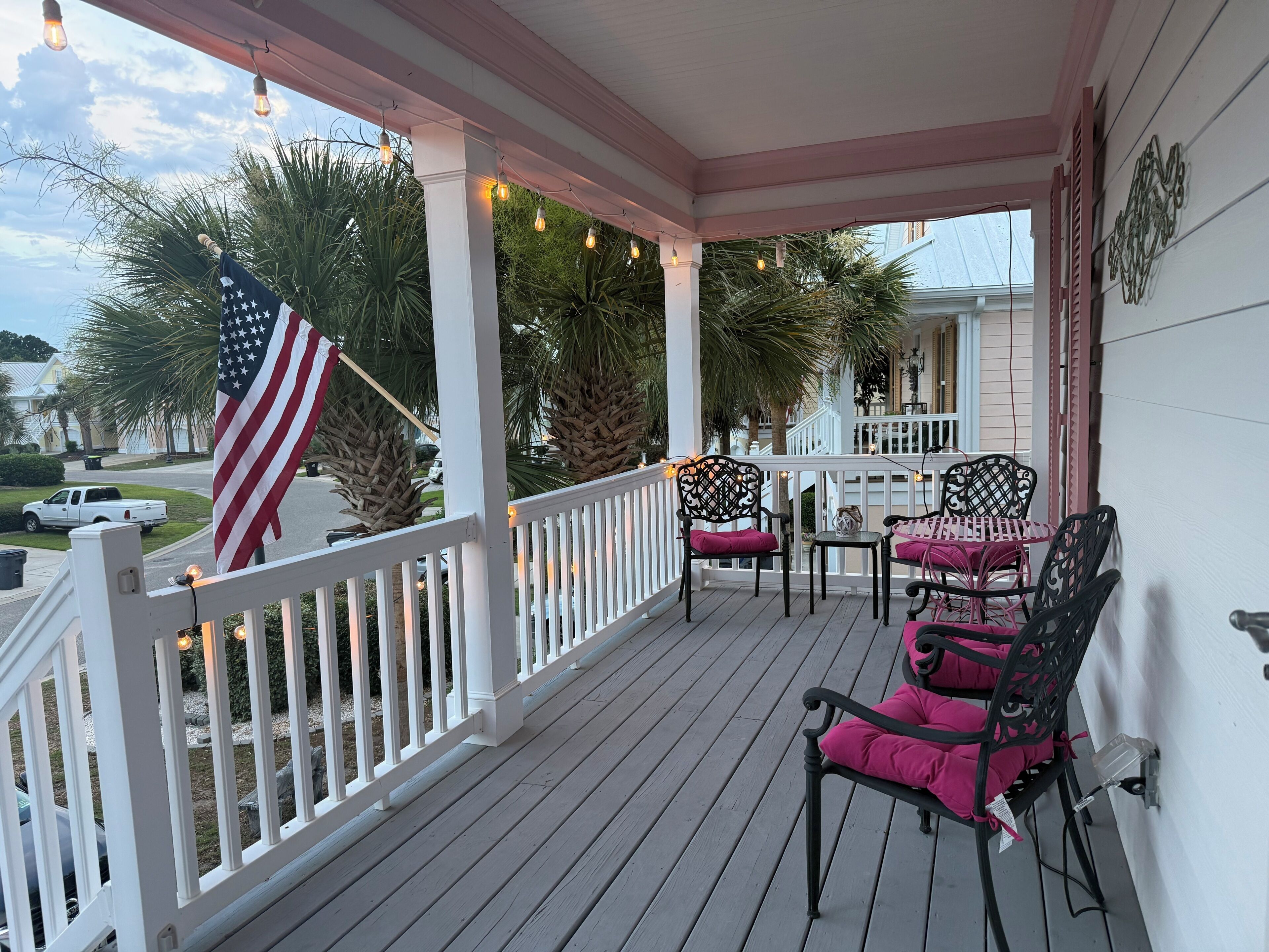 Een overdekt terras met een balustrade, buitenmeubels met kussens, een Amerikaanse vlag en sfeerverlichting.
