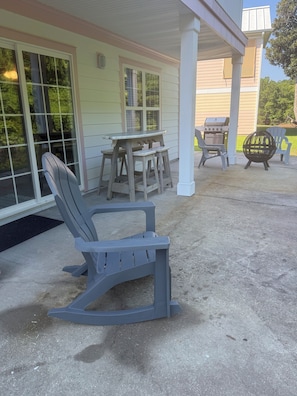 A patio with a rocking chair, a table, and two Adirondack chairs. - Pink Par-Tee Villa   Luxe Beach Getaway with Golf Cart (Murrells Inlet)