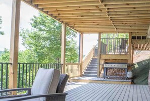 Eine Holzterrasse mit überdachtem Bereich, Gartenmöbeln, einem Kamin und einer Treppe, die zu einer oberen Ebene führt.
