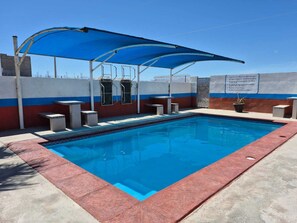 Ein rechteckiger Swimmingpool mit einer blauen Sonnensegelstruktur darüber, umgeben von einer niedrigen Mauer mit rotem und weißem Streifenmuster.