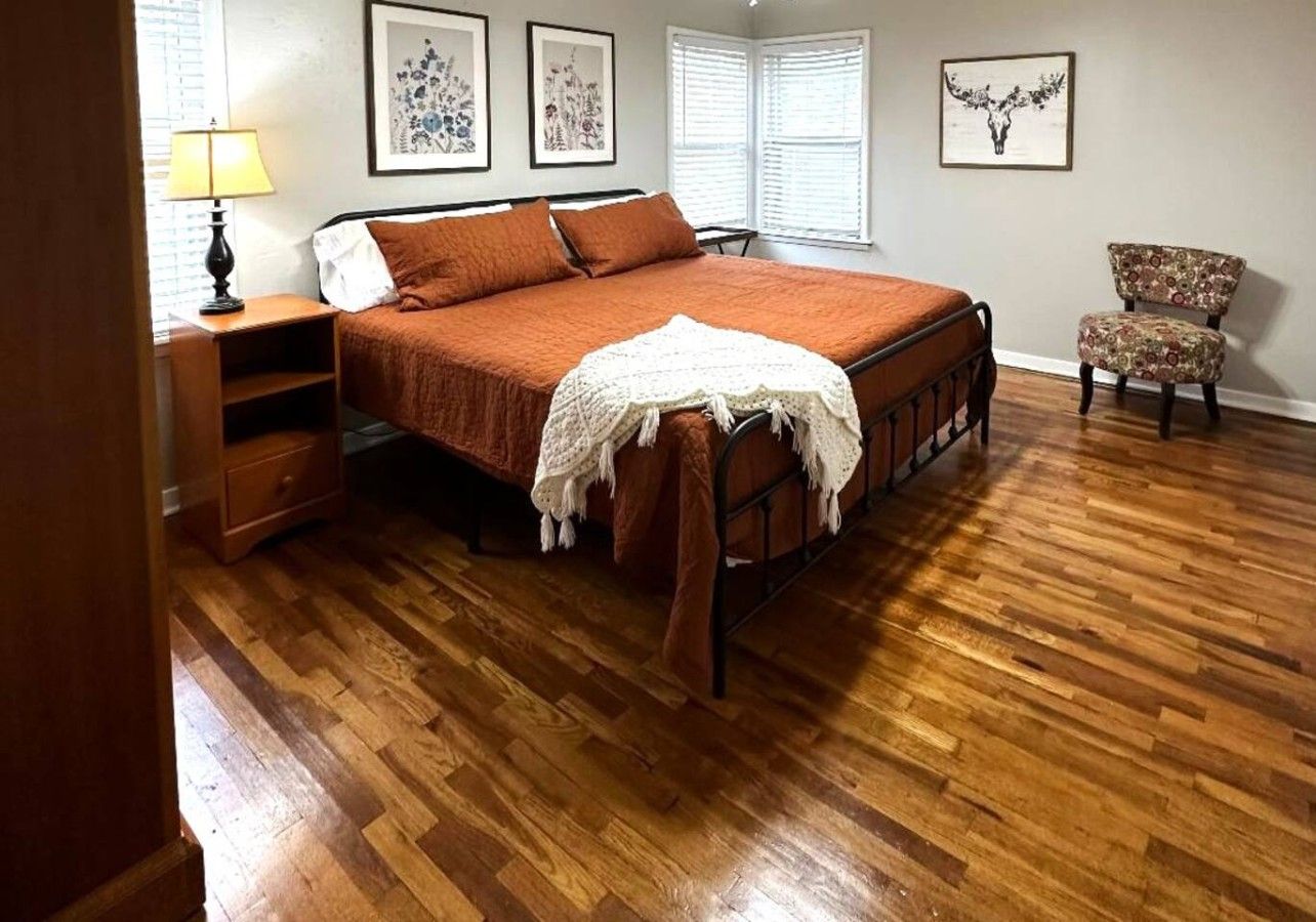 A bedroom with a wooden floor, a bed with a brown bedspread, a nightstand with a lamp, a chair, and framed artwork on the wall.