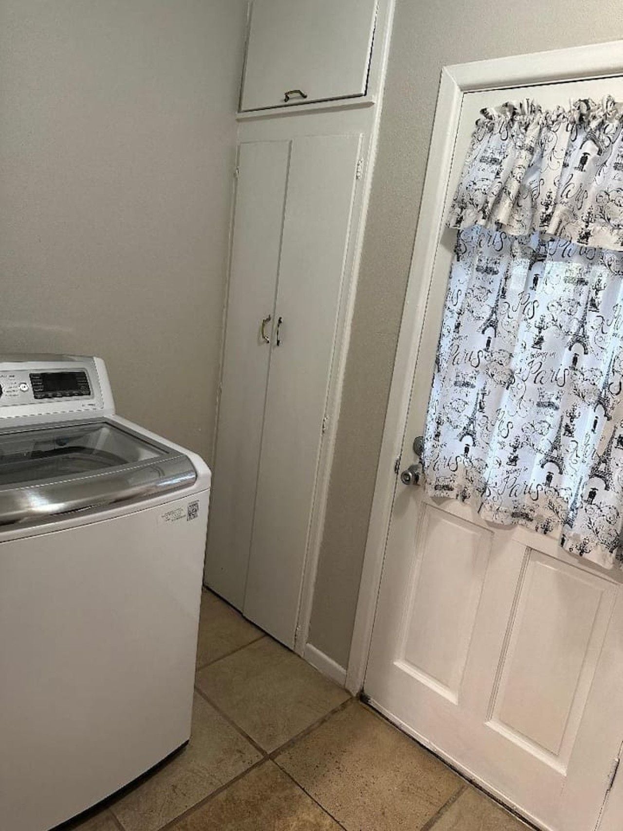Un lavadero con lavadora de carga frontal, un armario y una puerta con cortinas estampadas.