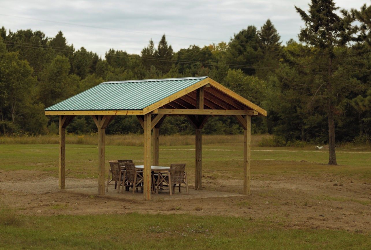 A wooden pavilion with a metal roof, surrounded by chairs and a table, set in a grassy field with trees in the background.