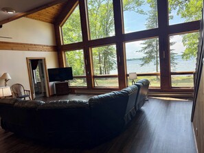 A spacious room with a large window, a leather sofa, a television, and a wooden floor. - Beautiful home, giant windows, sand beach, dock. Premier crystal clear lake. (Bemidji)
