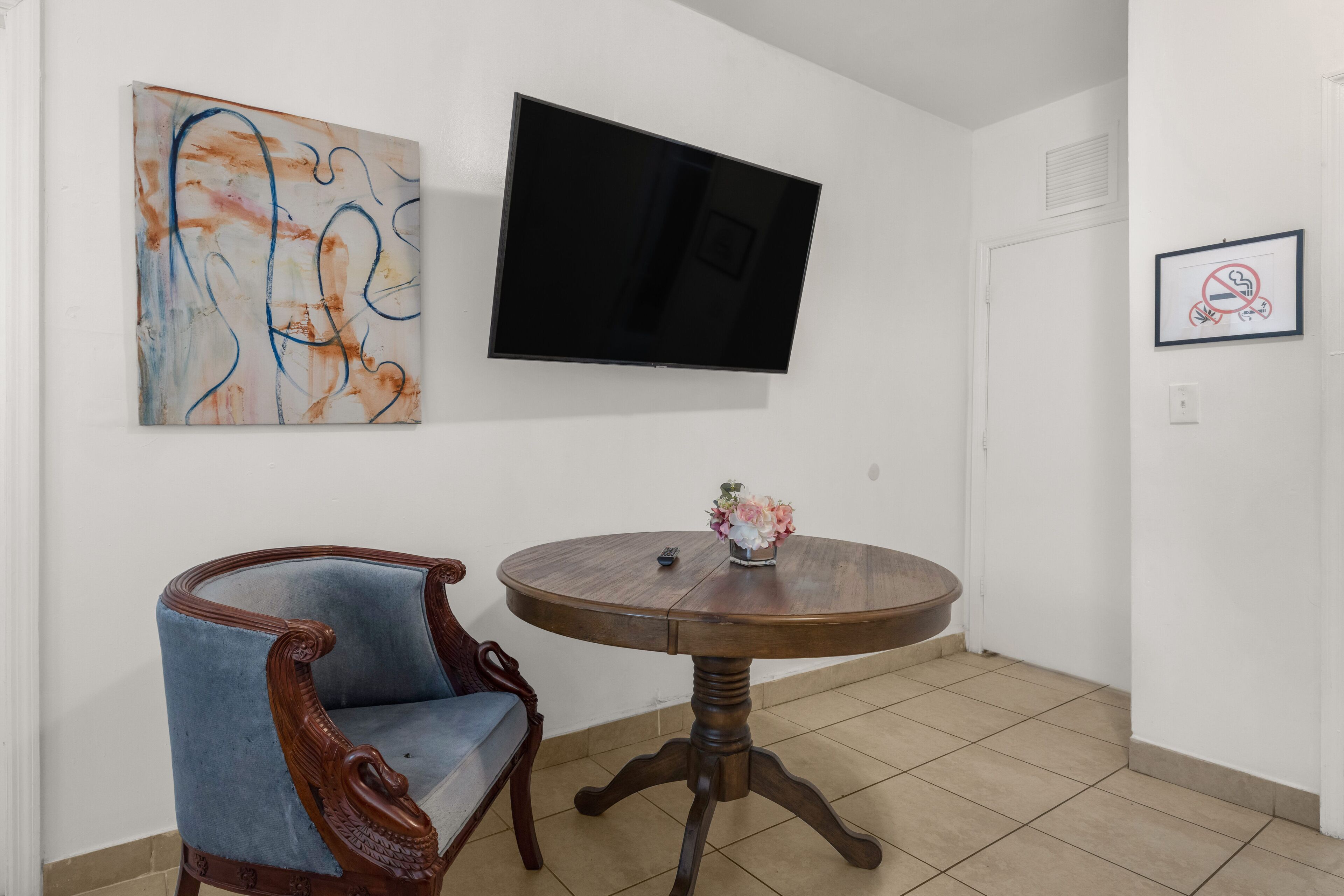 Une pièce avec un téléviseur fixé au mur, une table ronde en bois surmontée d’un vase de fleurs et un fauteuil bleu rembourré.