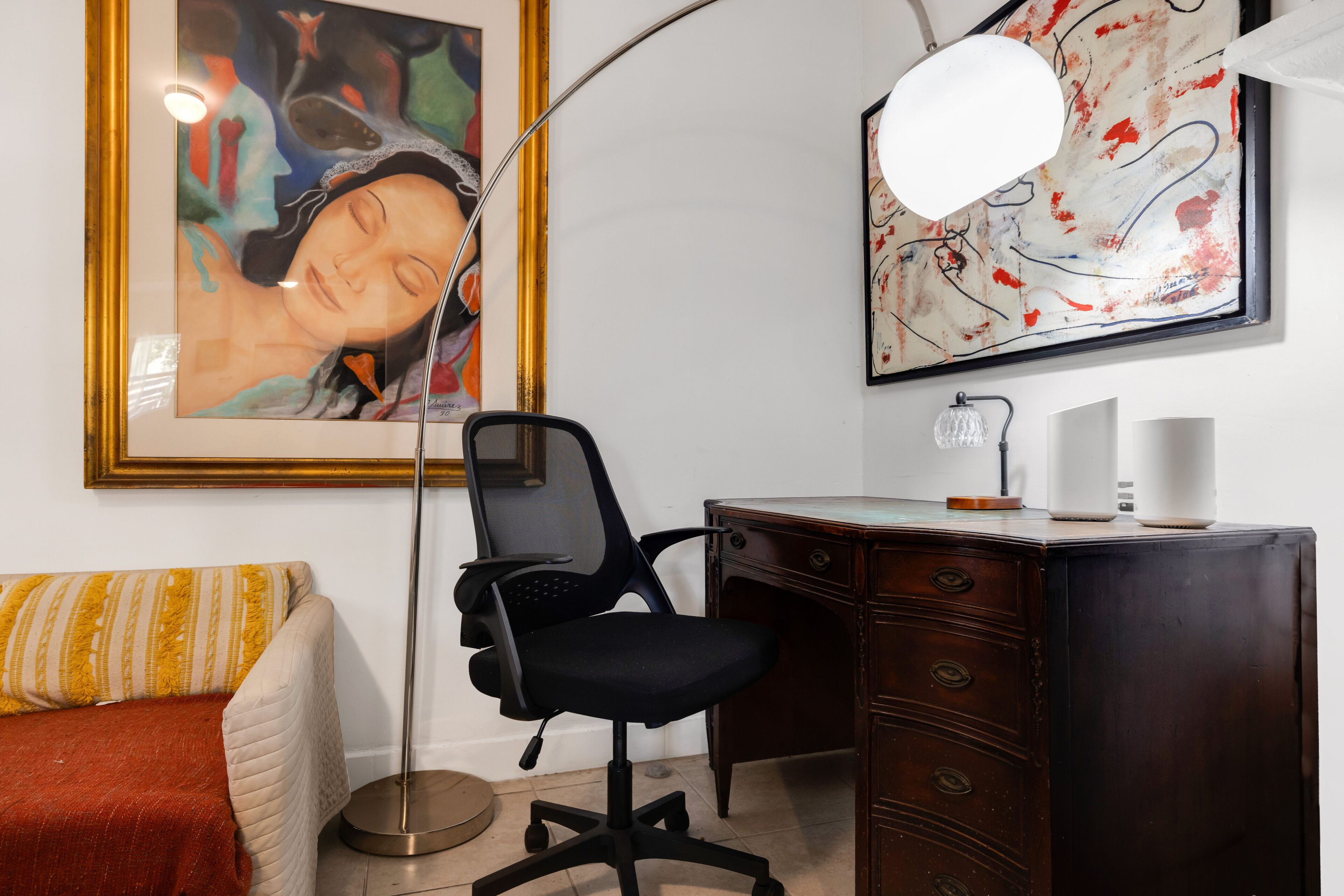 Ett rum med ett skrivbord, en stol, en lampa och konstverk på väggen.