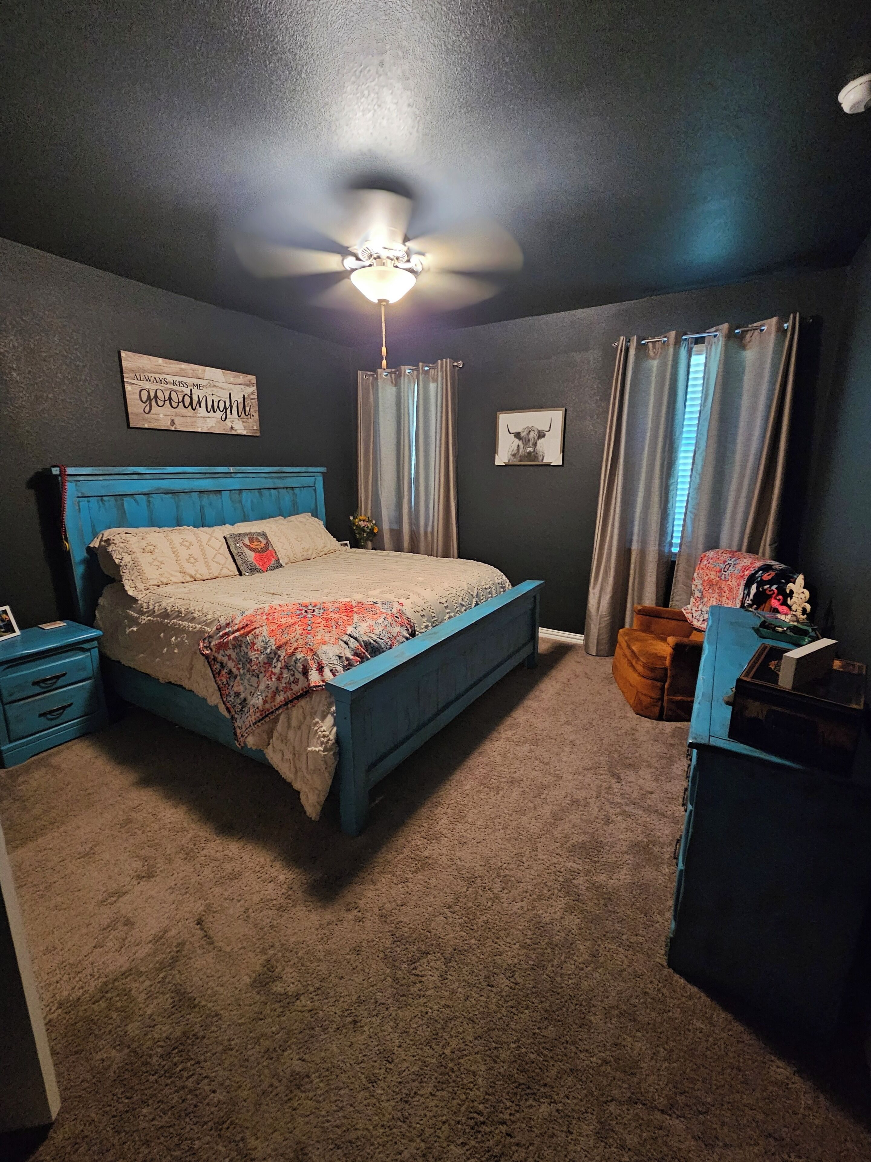 A bedroom with a bed, nightstands, a dresser, a ceiling fan, curtains, and a wall sign.