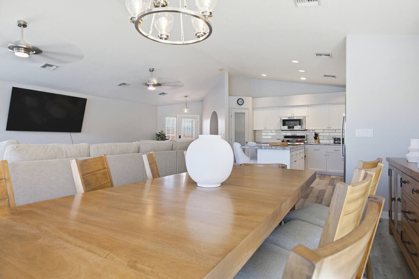 A spacious dining area with a long wooden table, cushioned seating, a modern chandelier, and a kitchen in the background.