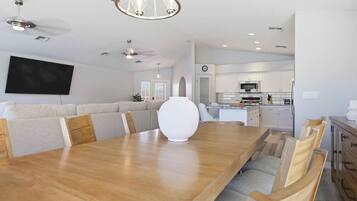 A spacious dining area with a long wooden table, cushioned seating, a modern chandelier, and a kitchen in the background.