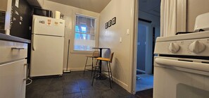 A compact kitchen with a refrigerator, microwave, and oven. A wicker chair is placed near the window, and a door leads to another room. - Charming 1920s Westbrae Berkeley Cottage with Soaking Tub! Walk to BART & Parks (Berkeley)