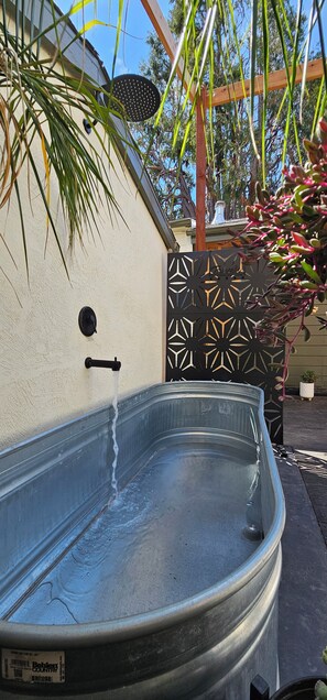 A metal hot tub with a label, a wall-mounted showerhead, and a decorative metal fence with geometric patterns. - Charming 1920s Westbrae Berkeley Cottage with Soaking Tub! Walk to BART & Parks (Berkeley)
