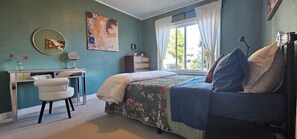 Une chambre à coucher avec un lit, un bureau, une chaise, une fenêtre avec des rideaux et un tableau accroché au mur.