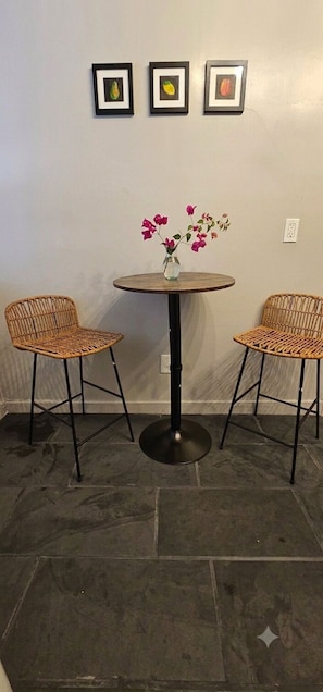 A round table with two wicker chairs, a vase with pink flowers, and three framed pictures on the wall. - Charming 1920s Westbrae Berkeley Cottage with Soaking Tub! Walk to BART & Parks (Berkeley)
