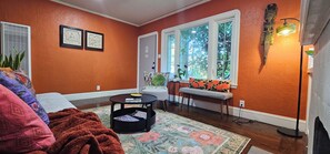 A living room with orange walls, a sofa, a coffee table, and a window with a view of trees. - Charming 1920s Westbrae Berkeley Cottage with Soaking Tub! Walk to BART & Parks (Berkeley)