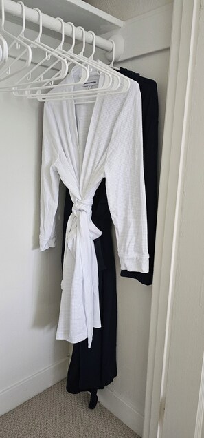A white robe tied at the waist hanging on a white metal drying rack in a corner of a room with white walls and a carpeted floor.