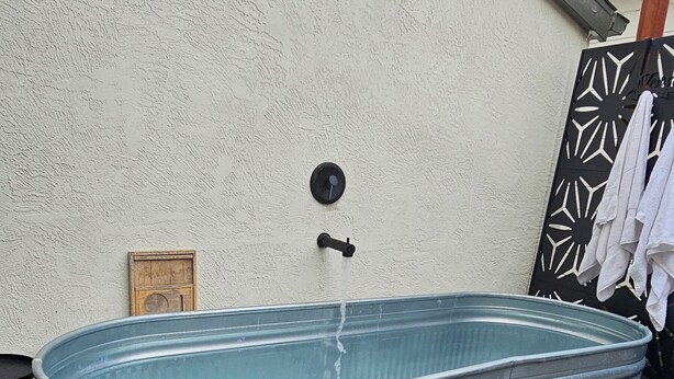 A metal hot tub in an outdoor setting with a wooden deck, a white wall, and a black and white patterned gate. - Charming 1920s Westbrae Berkeley Cottage with Soaking Tub! Walk to BART & Parks (Berkeley)