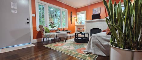 A living room with a sofa, armchair, and a coffee table. There is a fireplace with a mantel, and a large window with a view of trees.
