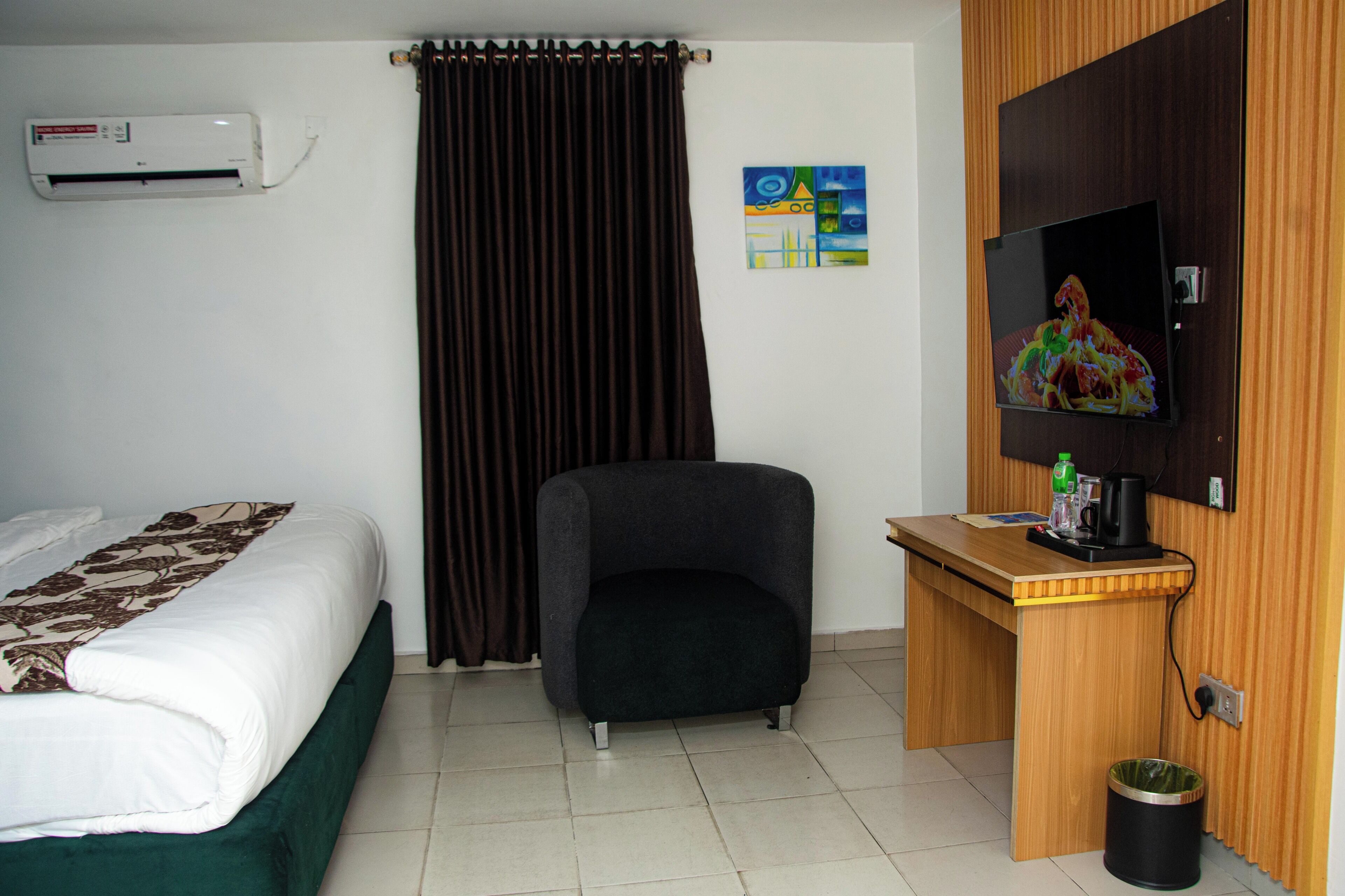 A hotel room with a bed, a desk, a chair, an air conditioner, a television, and a painting on the wall.