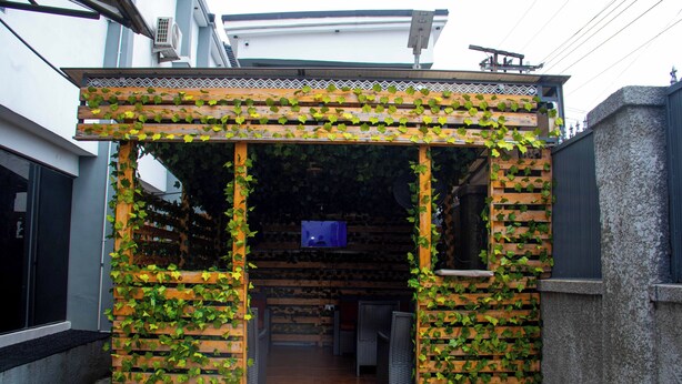 Un espacio de estar al aire libre cubierto, con listones de madera y vegetación, una pantalla de televisión y vista al interior.