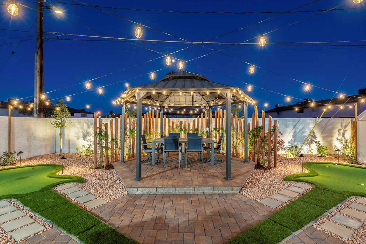 A gazebo with a table and chairs, surrounded by a garden with a putting green.