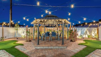 A gazebo with a table and chairs, surrounded by a garden with a putting green.