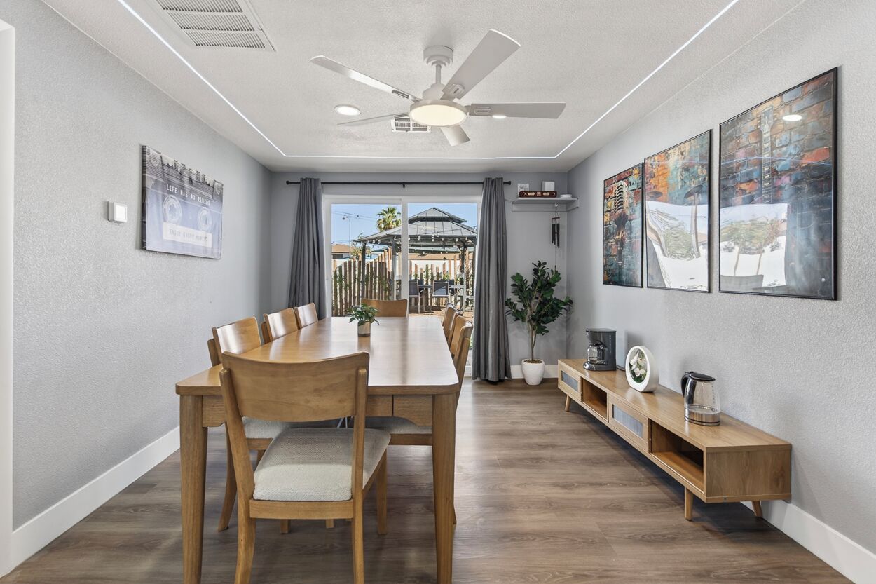 A dining area with a long wooden table, chairs, a ceiling fan, and a view of a building through a glass door.