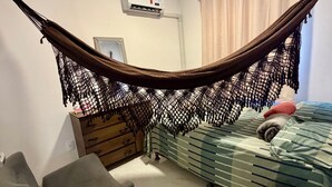 A bedroom with a hammock, a bed with striped bedding, a chair, a dresser, and an air conditioning unit. - Ceará Corner - Apartment on Futuro Beach (Fortaleza)