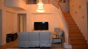 Un salon avec une grande télévision, un lustre, un escalier et un canapé.